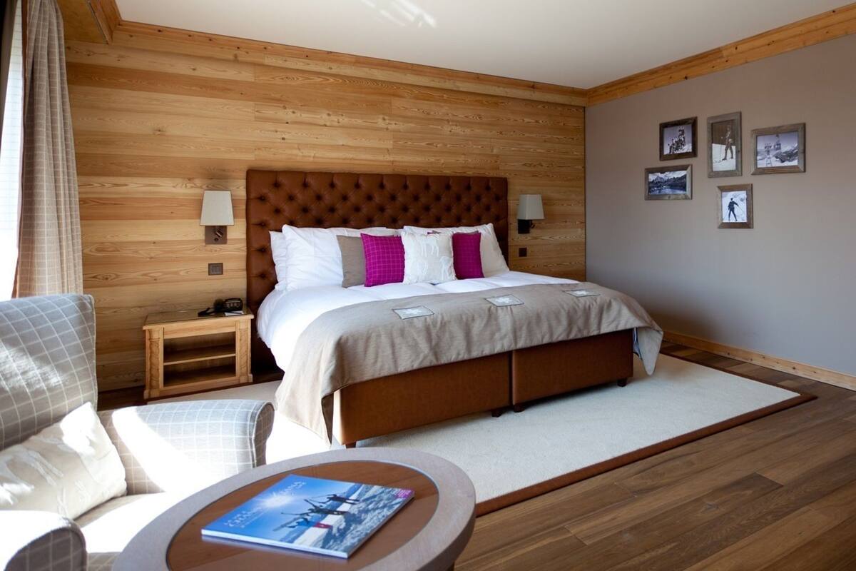 A bedroom with a wooden headboard, a bed with white and pink pillows, a bedside table with a lamp, a chair, and a small table with a book.