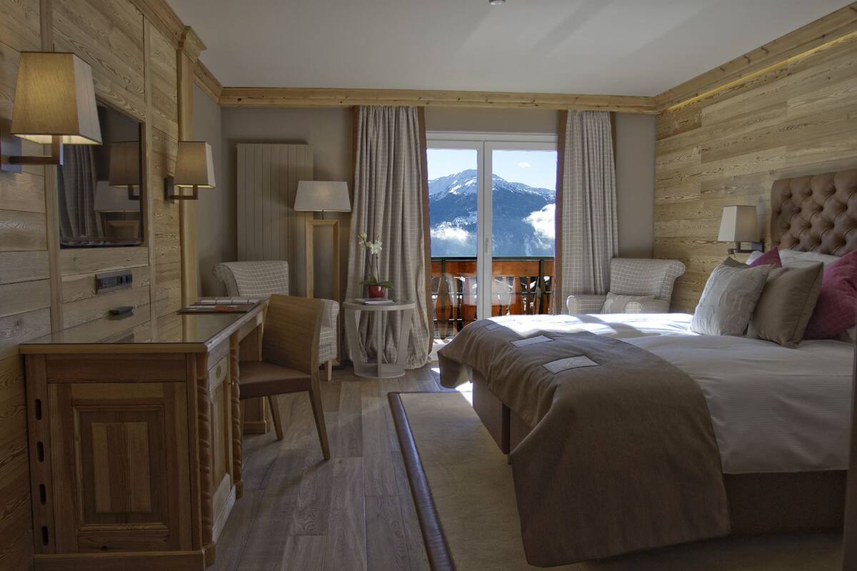 A hotel room with a bed, a desk, a chair, a small table, and a view of mountains through the window.