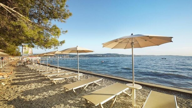 A beachside area with sun loungers and umbrellas, a pebble beach, and a view of the sea and distant hills. - Villas with Balcony or Terrace - Hotel & Resort Adria Ankaran (Ankaran)