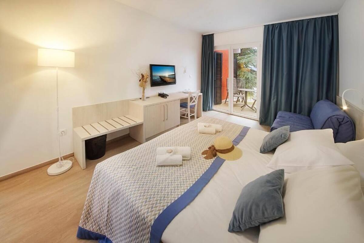 Quarto de hotel com cama, escrivaninha, cadeira, televisão e varanda com cortinas.