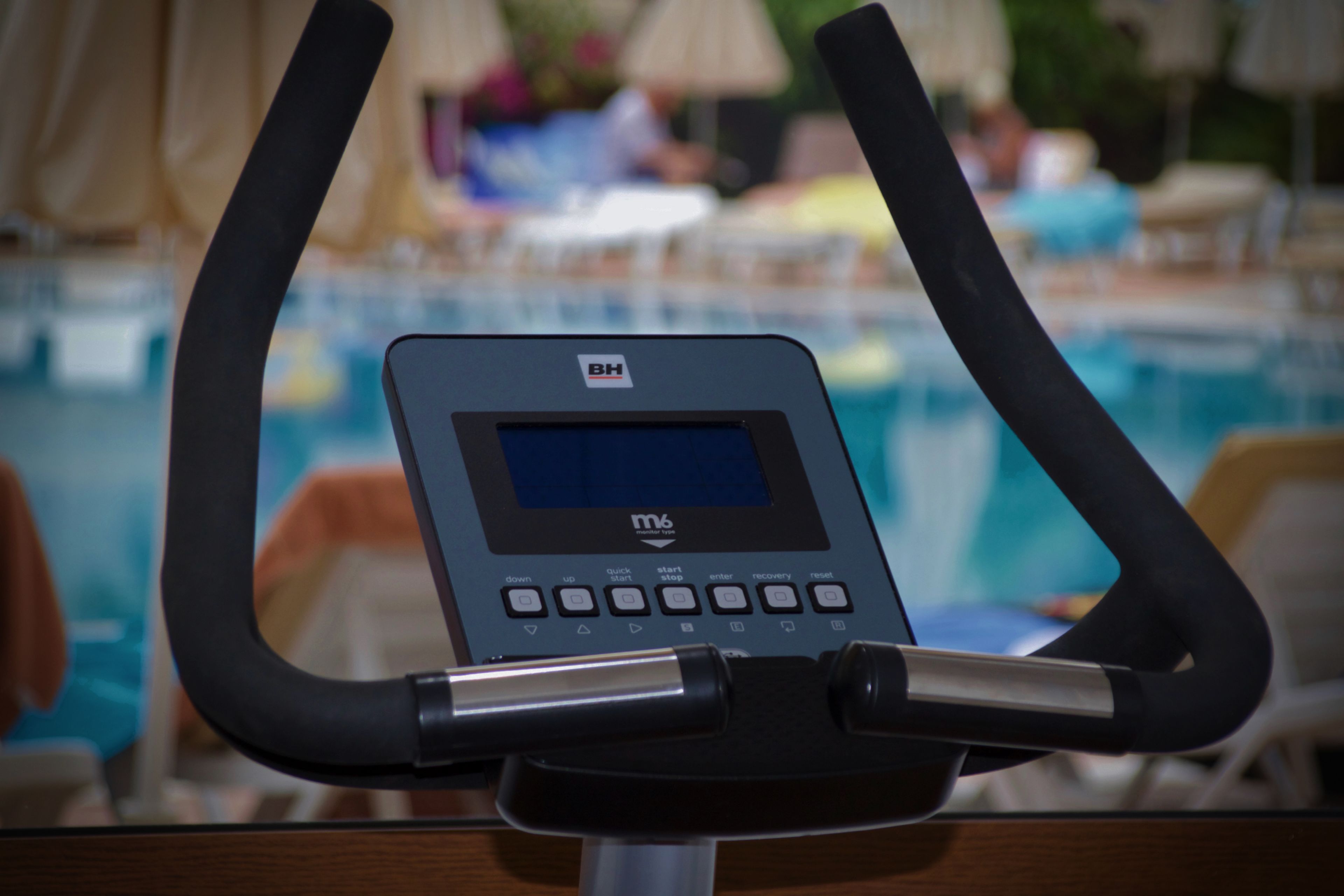 Una máquina de ejercicio de m6 con pantalla digital, ubicada cerca de una zona de piscina con sillones de jardín y sombrillas.