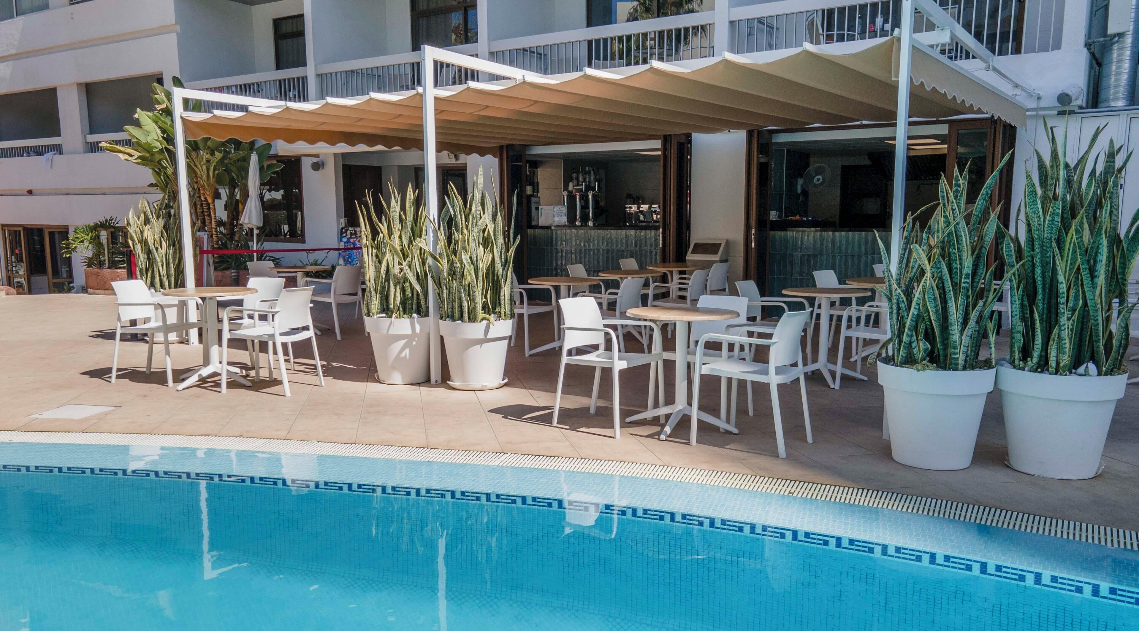 Un área de piscina al aire libre con sillas y mesas blancas, rodeada de plantas en macetas.