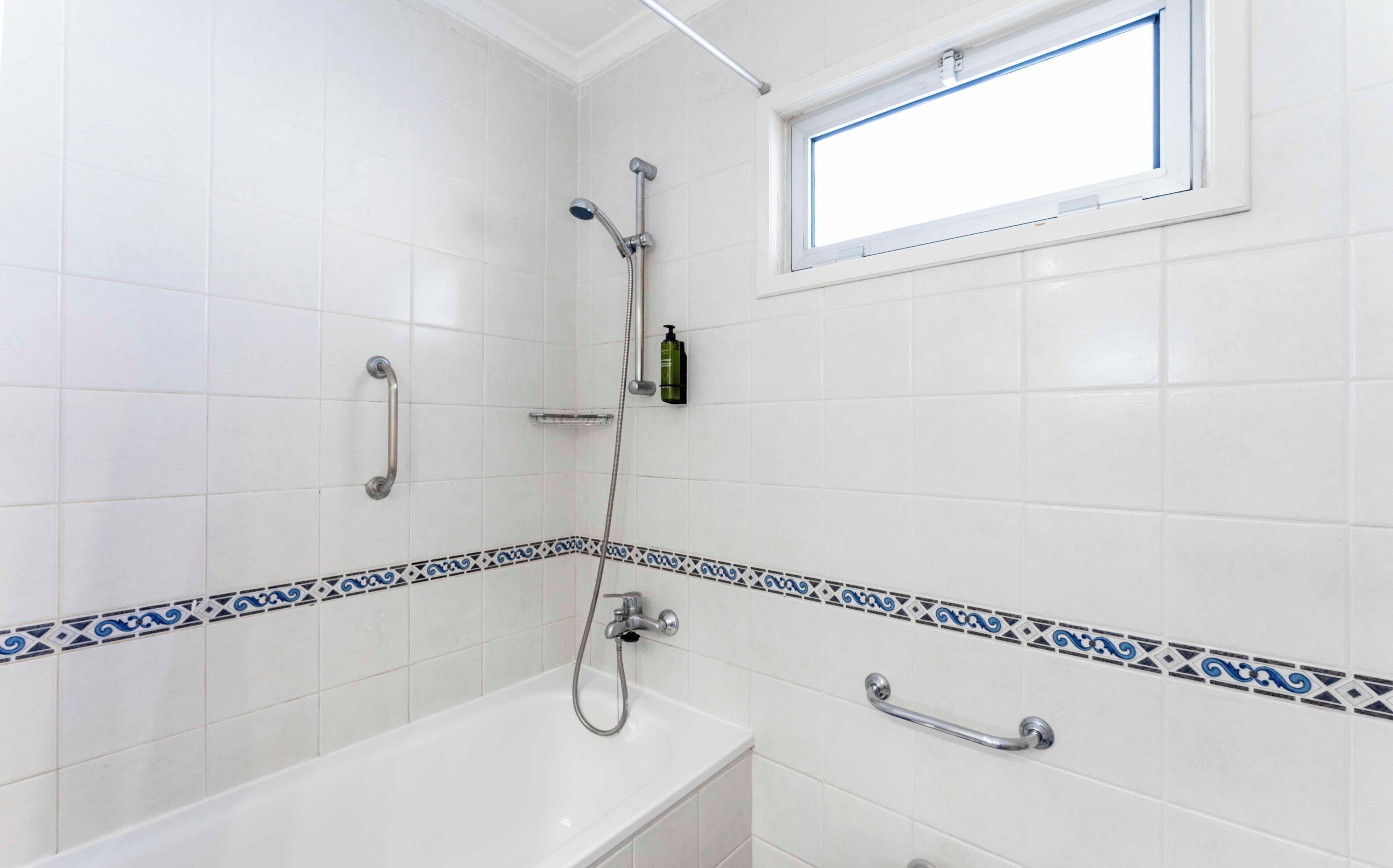 Un baño con paredes de azulejos blancos, una bañera, una ducha y un ventanal.