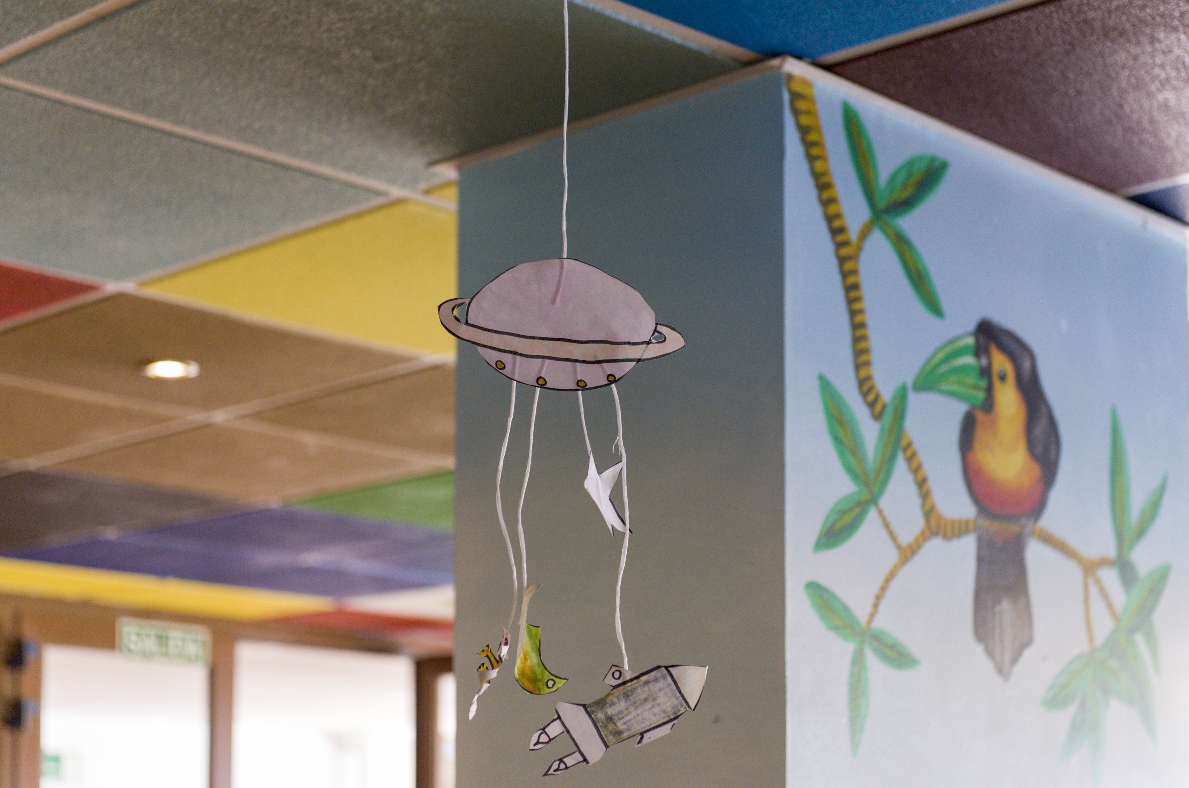 Een plafond met een kleurrijk geometrisch patroon en een decoratieve hanglamp met een vogel en een raket.