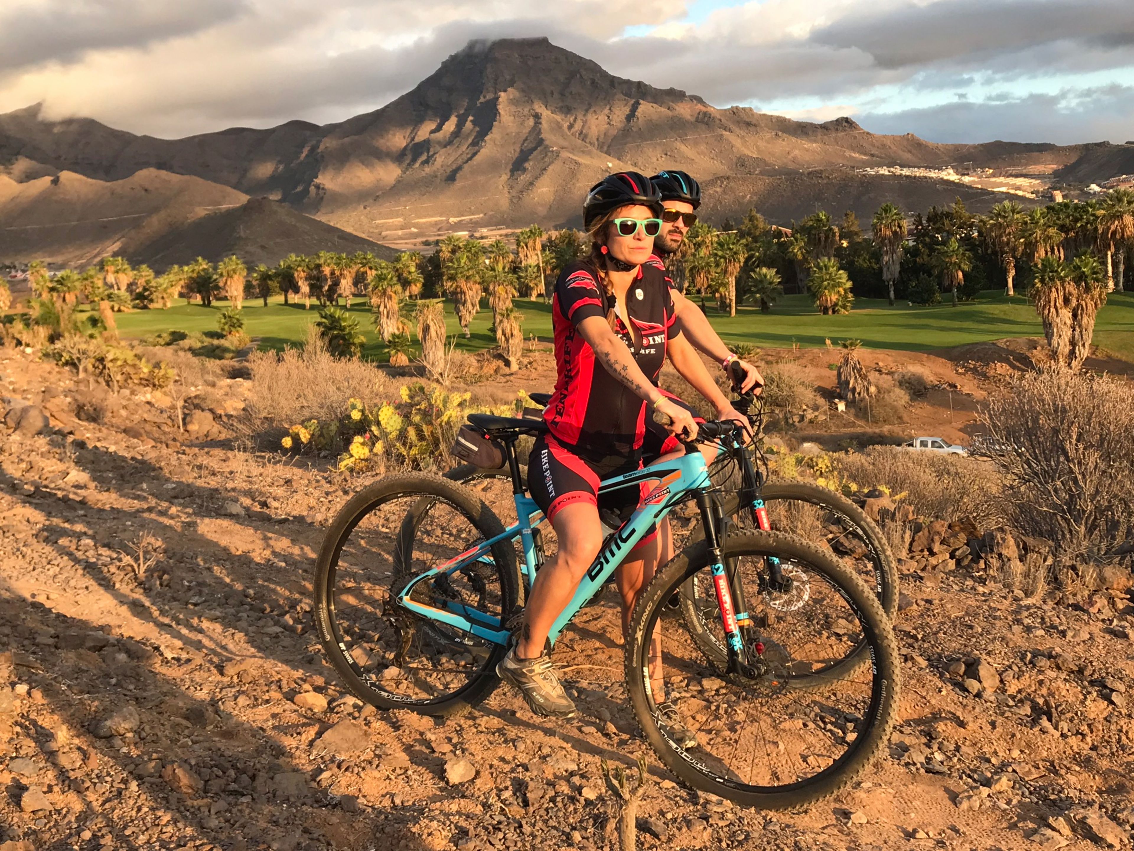 Dos personas en un sendero de bicicleta de montaña, con montañas al fondo.