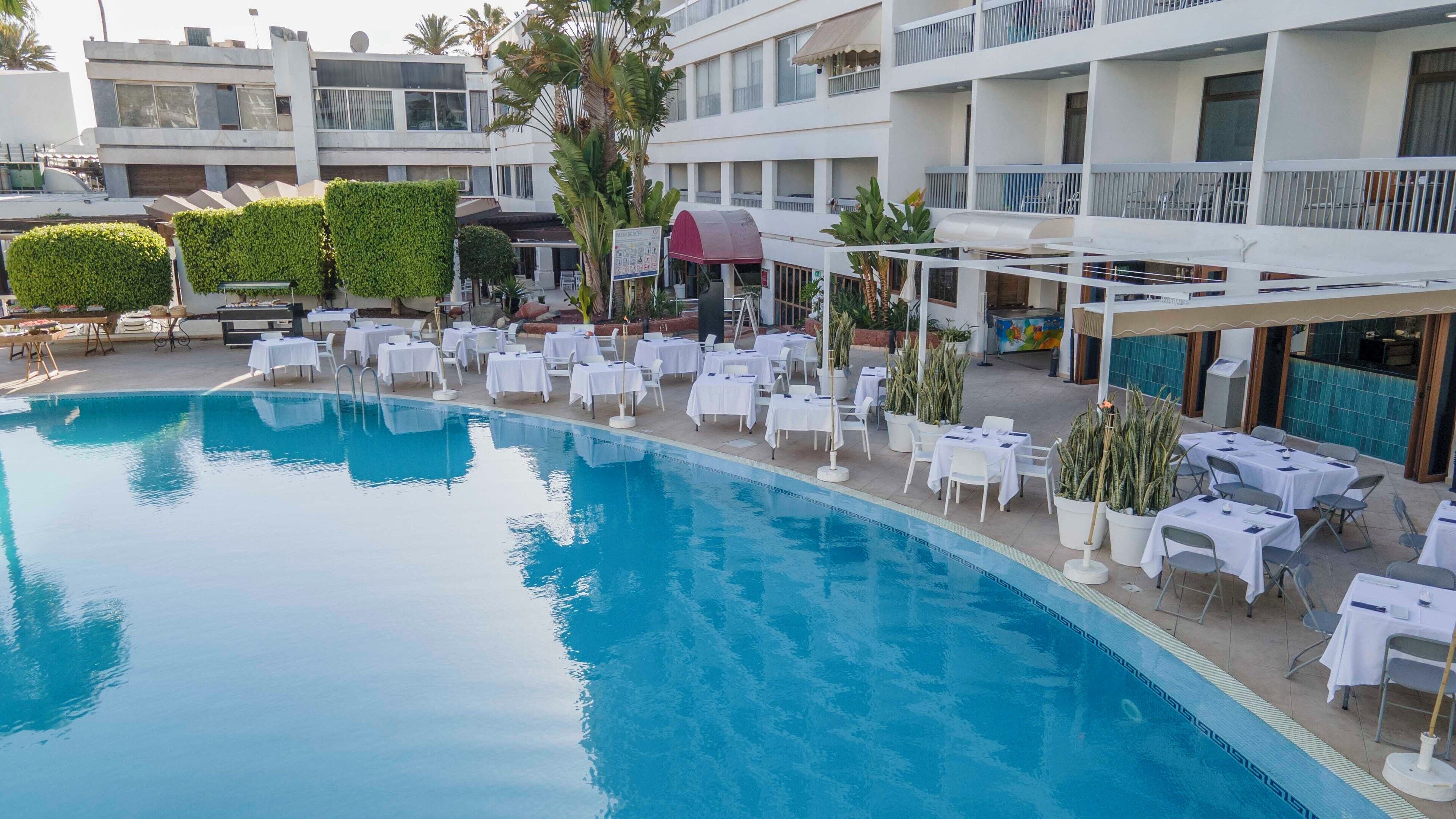 Ein Poolbereich mit Tischen und Stühlen, eine überdachte Lounge-Ecke und im Hintergrund ein Gebäude mit Balkonen.
