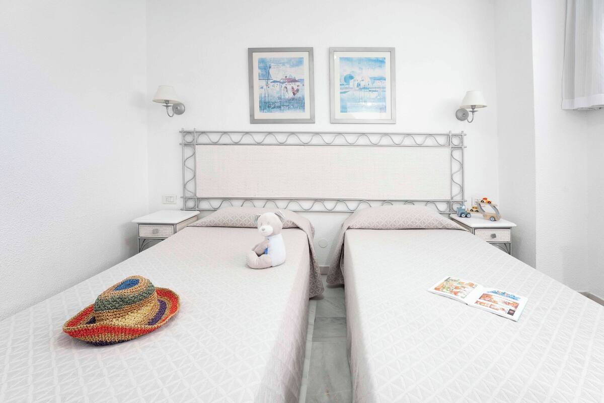 A hotel room with two beds, a nightstand with a lamp, a wall-mounted shelf with decorative items, and two framed pictures on the wall.