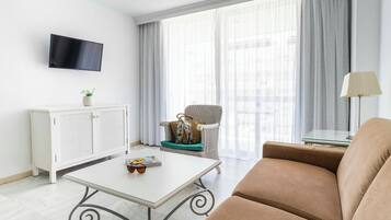 Ein modernes Wohnzimmer mit einem weißen Sofa, einem weißen Couchtisch mit Sonnenbrille und Zeitschrift, einem Korbstuhl mit einer Handtasche, einer weißen Kommode, einem an der Wand befestigten Fernseher und einem großen Fenster mit leichtem Vorhang.