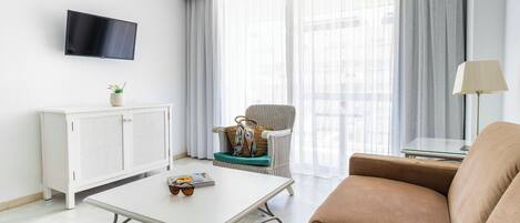 Un salon moderne avec un canapé blanc, une table basse blanche agrémentée de lunettes de soleil et d’un magazine, un fauteuil en osier avec un sac à main, une armoire blanche, un téléviseur fixé au mur et une grande fenêtre avec des rideaux légers.
