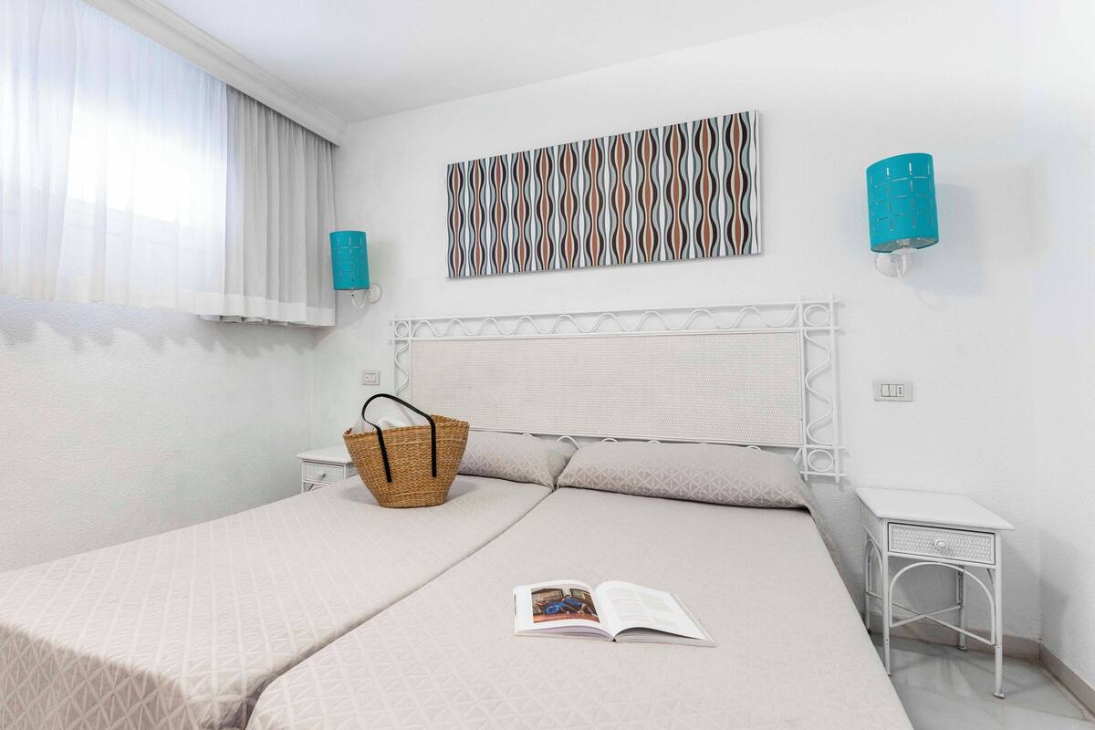 A neatly arranged bedroom with a bed, a bedside table, a wall-mounted lamp, and a window with sheer curtains.