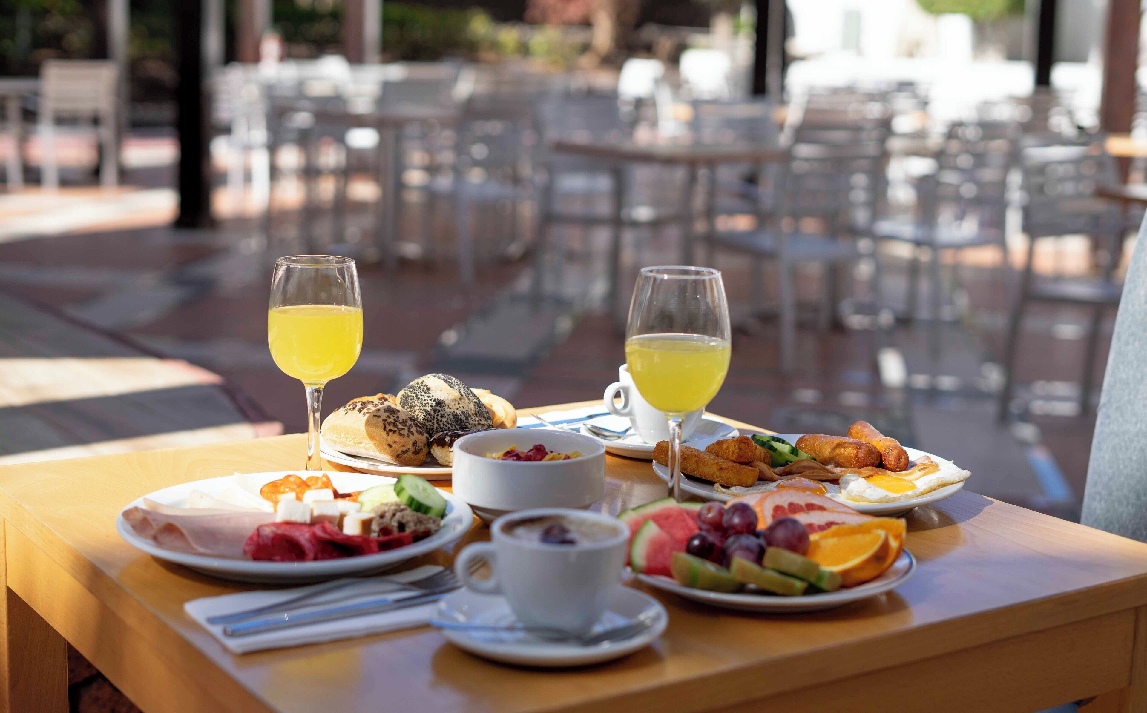 A table set with breakfast items including pastries, fruit, and beverages, with outdoor seating in the background.