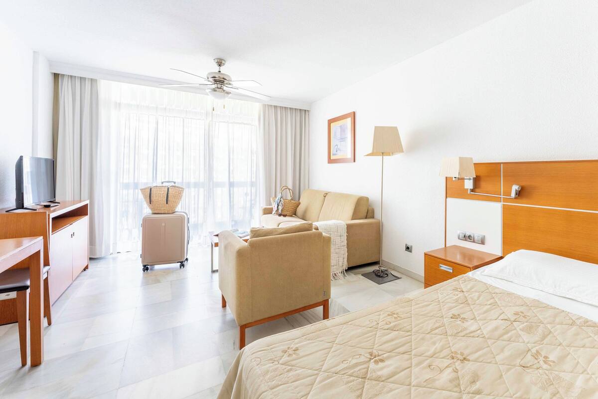 A hotel room with a bed, a desk with a TV, a chair, a sofa, a ceiling fan, and a window with curtains.