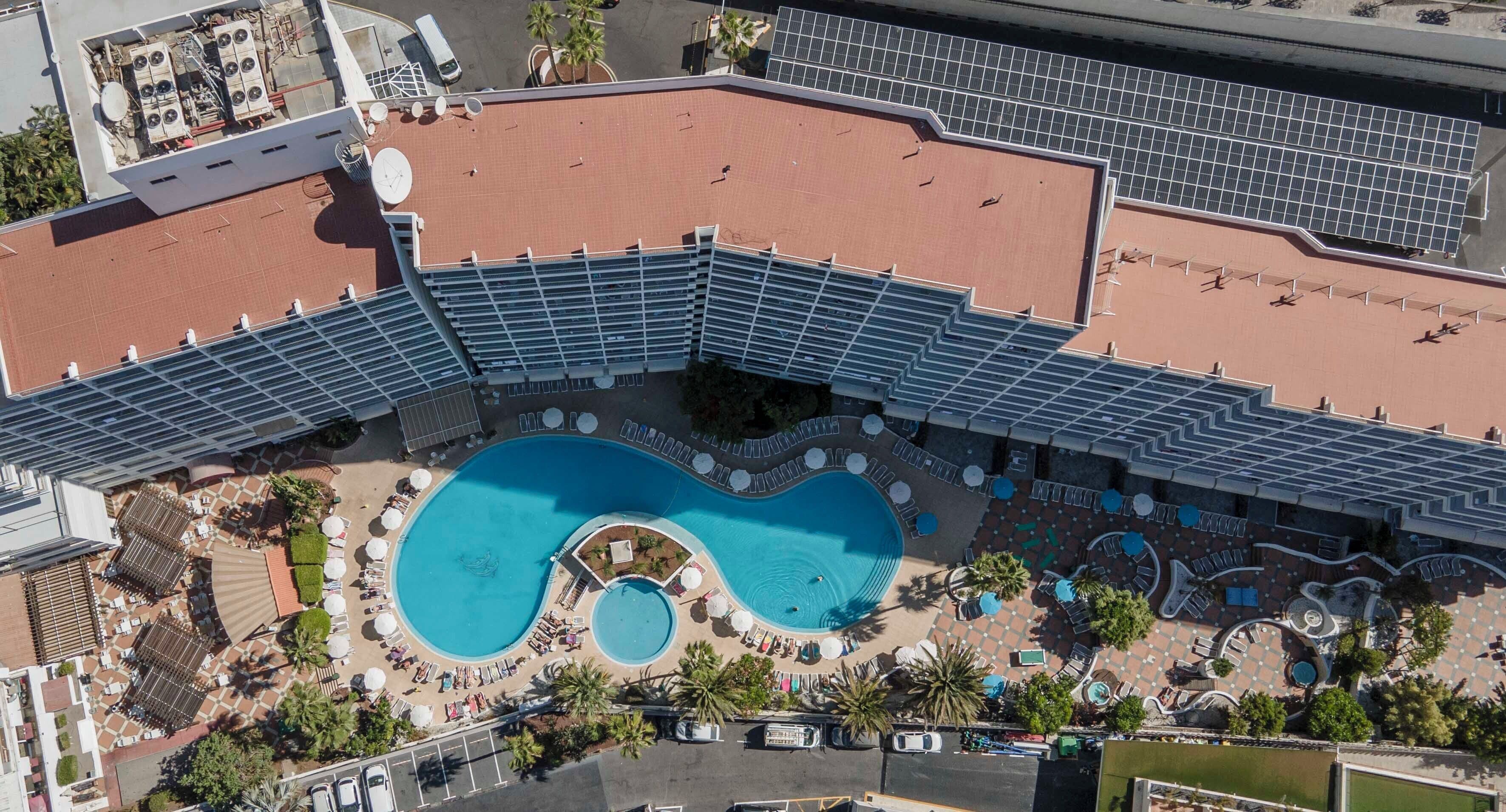 An aerial view of a hotel with a large swimming pool, sun loungers, and a covered area with a curved roof.