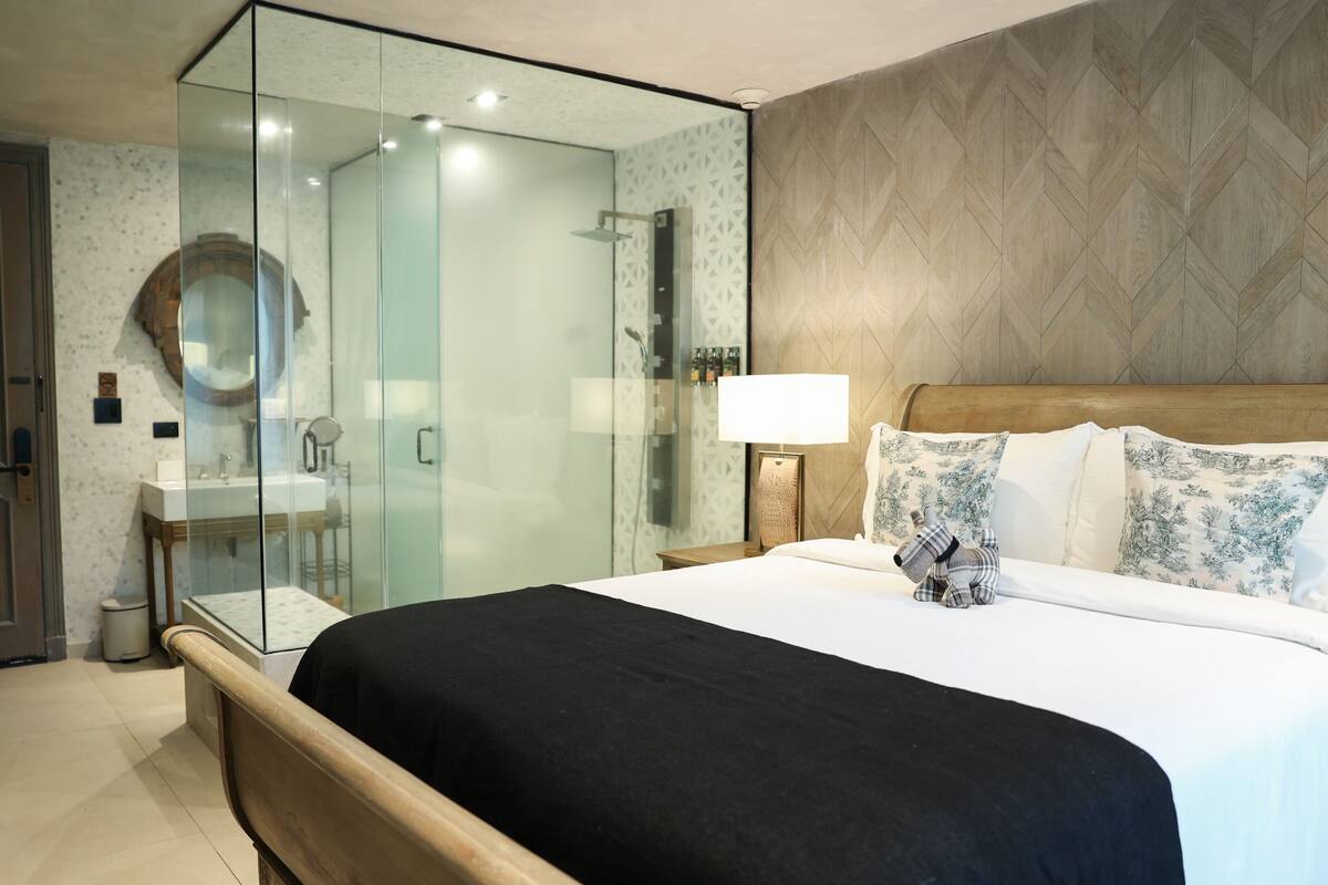 A modern hotel room with a glass-enclosed shower, a wooden headboard, and a neatly made bed with a black bedspread.