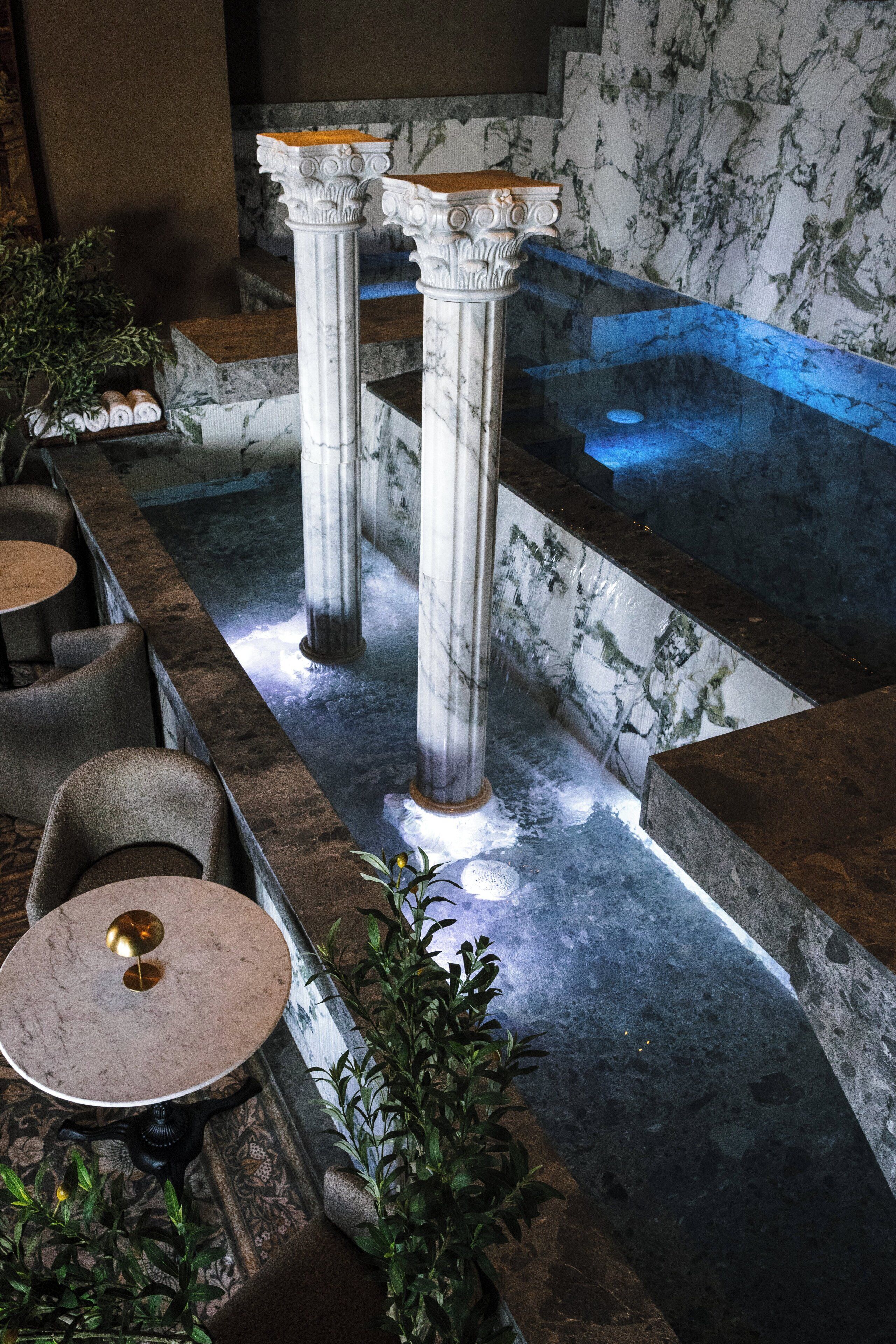 Un área de spa con columnas de mármol, una bañera de hidromasaje y asientos con mesas y sillas.
