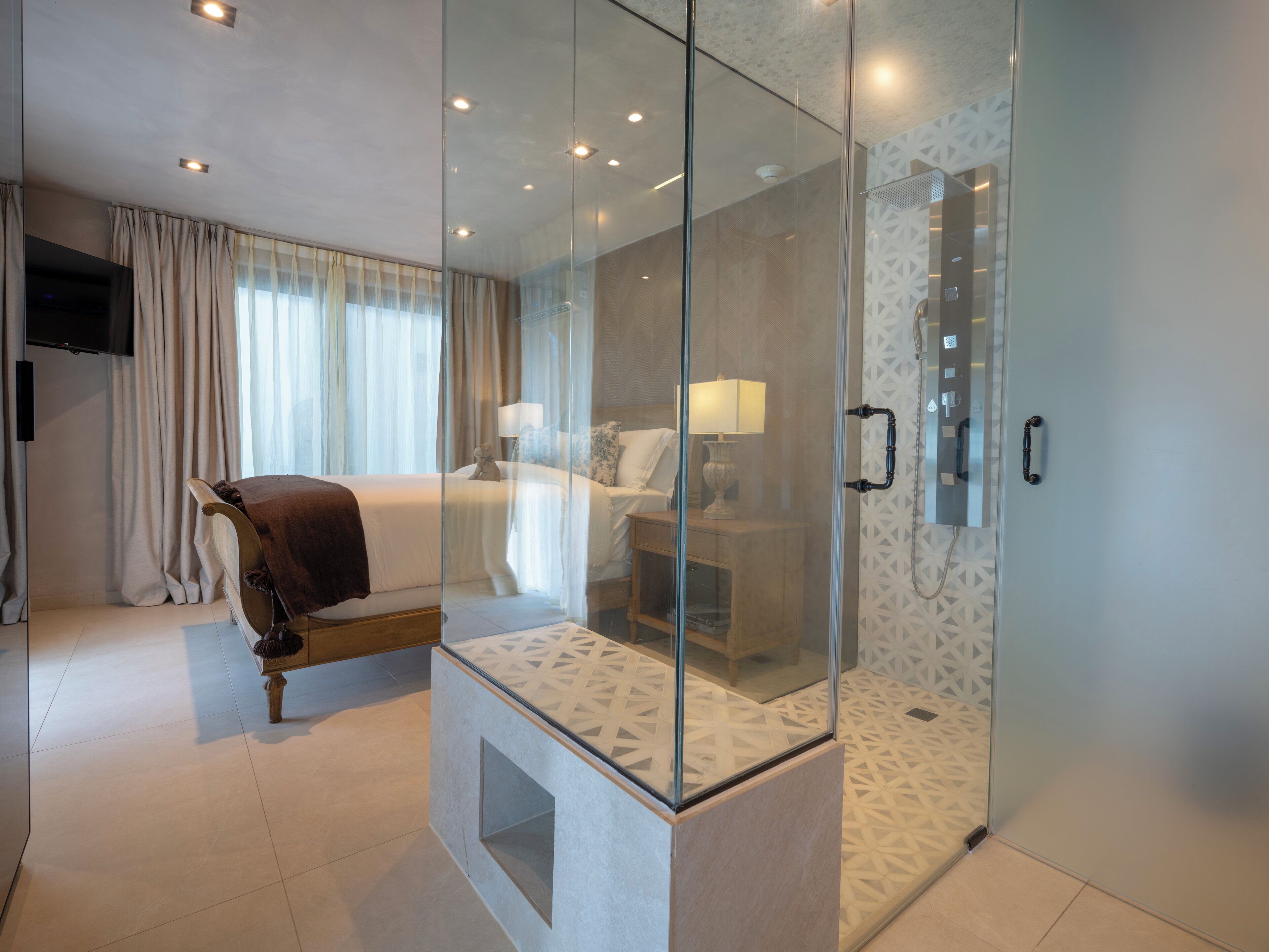Una habitación de hotel moderna con ducha envuelta en vidrio, cama con cabecero de madera y mesita de noche con lámpara.