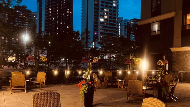 An outdoor seating area with wicker chairs and tables, a potted plant with flowers, and city buildings in the background. - Staybridge Suites Hamilton Downtown by IHG (Hamilton)
