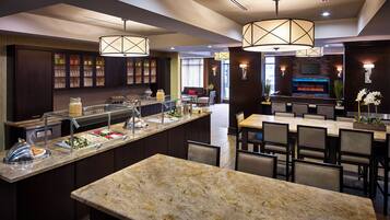 A modern hotel lobby with a central bar area, seating arrangements, and a buffet counter.
