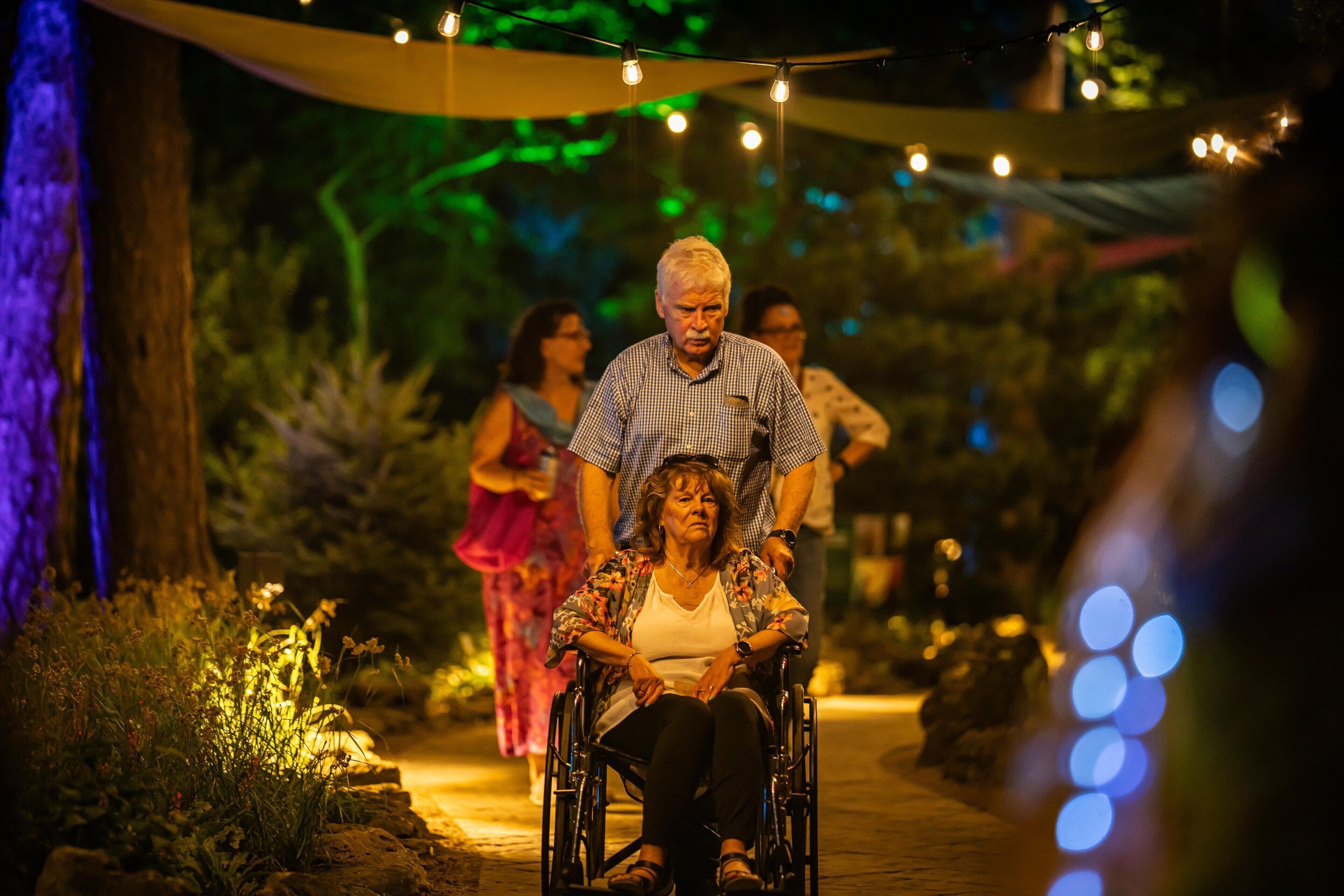 A pathway with string lights, a wheelchair, and people walking.