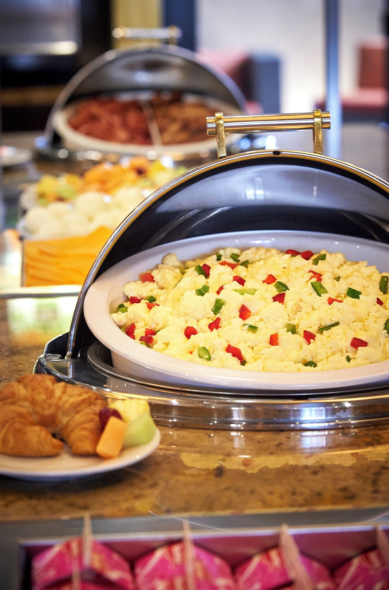 A buffet with various dishes, including scrambled eggs with vegetables, covered dishes, and a plate with a croissant.