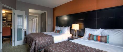 A hotel room with two beds, a headboard, a nightstand, a lamp, and a bathroom visible through an open door.