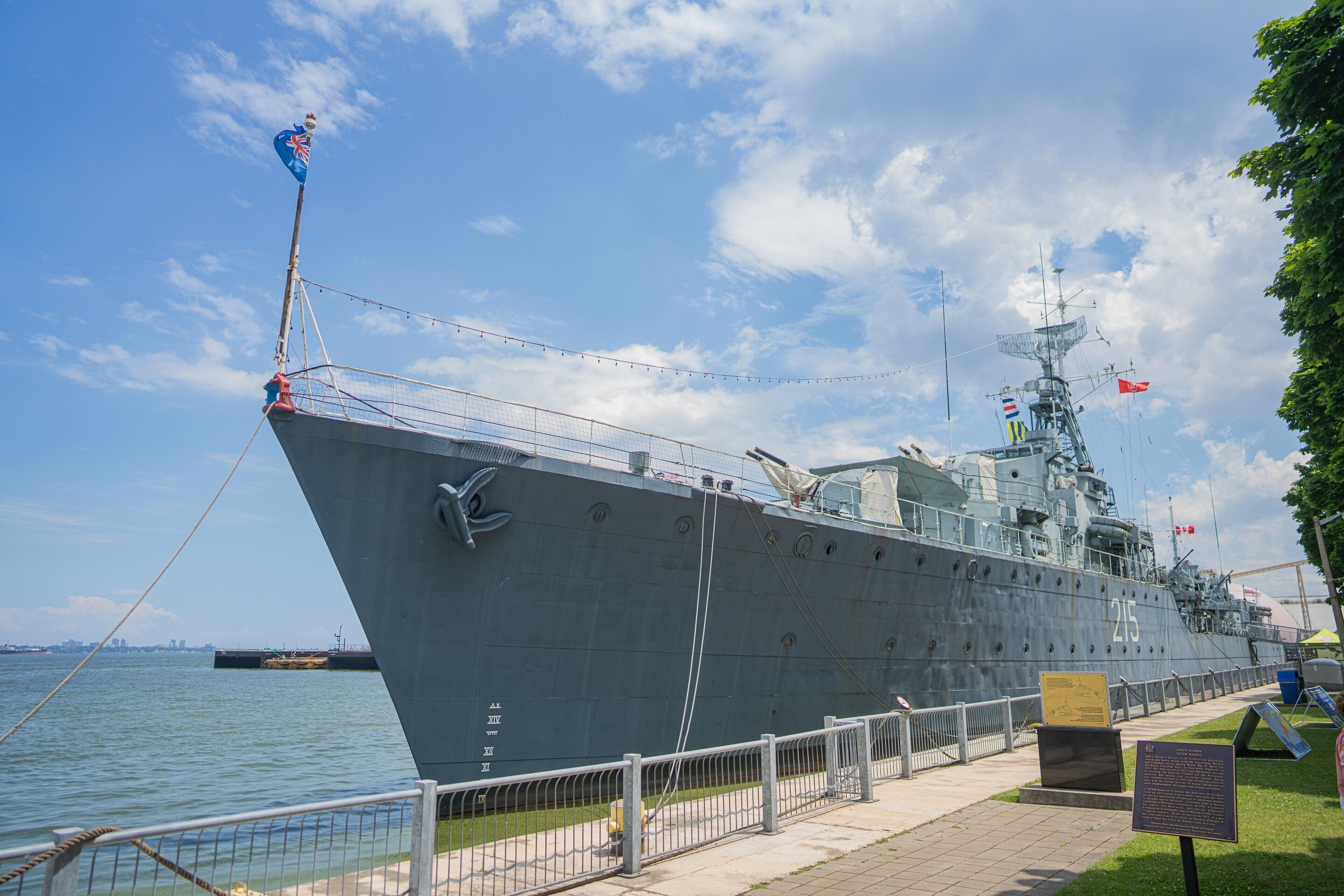 A large naval ship docked at a port with a clear sky and a grassy area in the foreground.