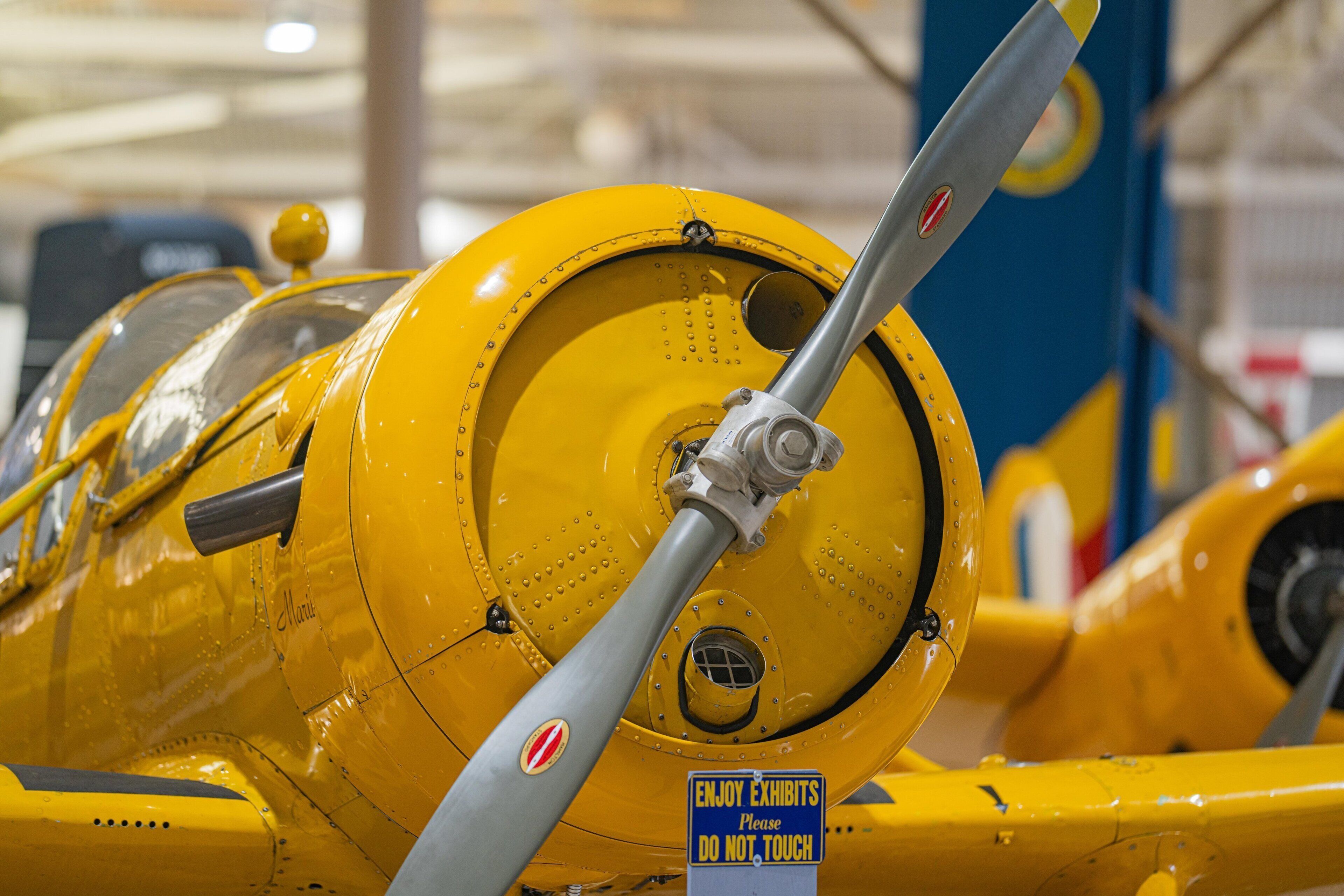 Un avión amarillo con hélice, un letrero que dice "DISFRUTA LAS EXHIBICIONES Por favor, NO TOCAR" y un fondo azul y amarillo.