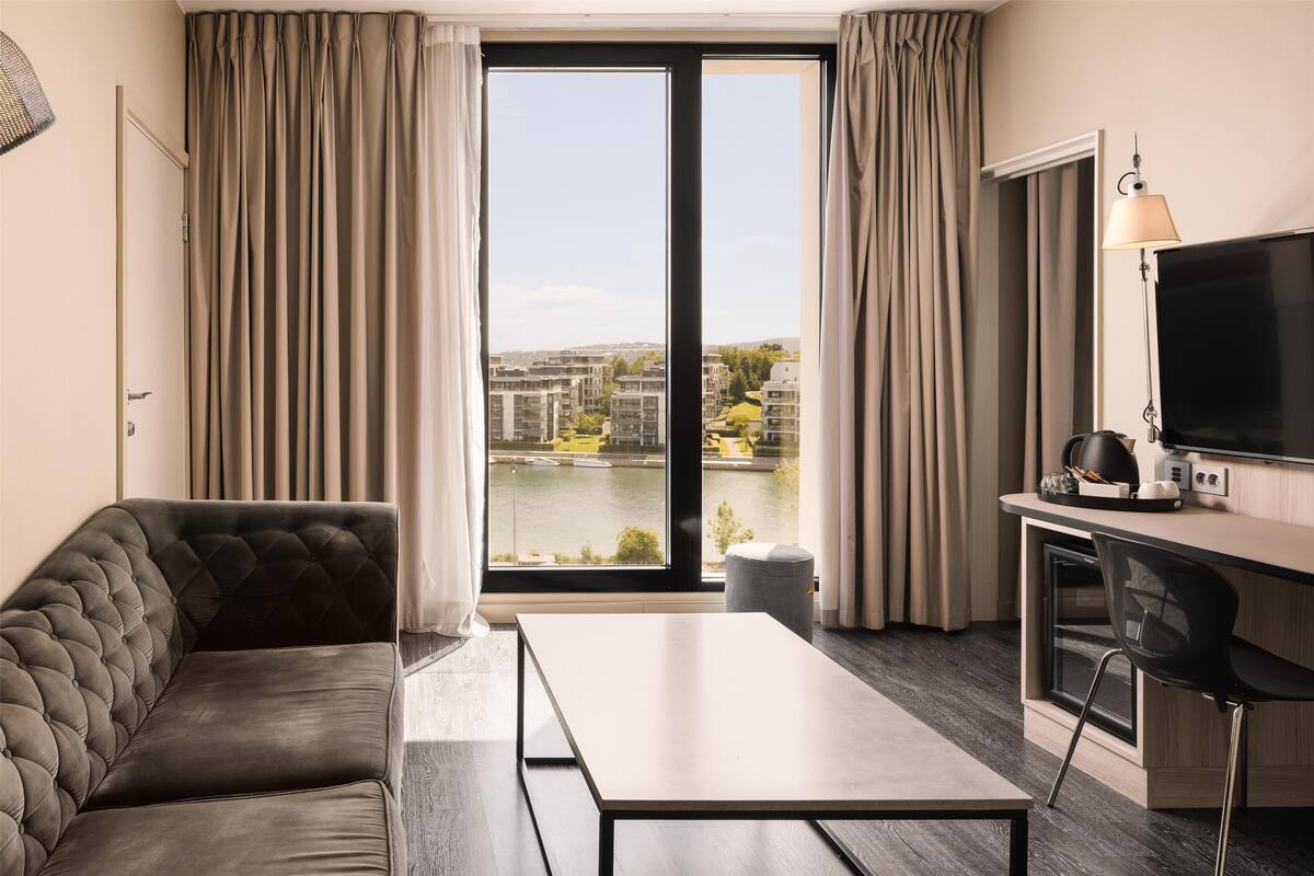 Una habitación de hotel moderna con un ventanal grande, un sofá, un escritorio y un televisor.