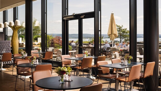 An outdoor dining area with tables and chairs, large windows, and a view of the water. - Scandic Fornebu (Bærum)