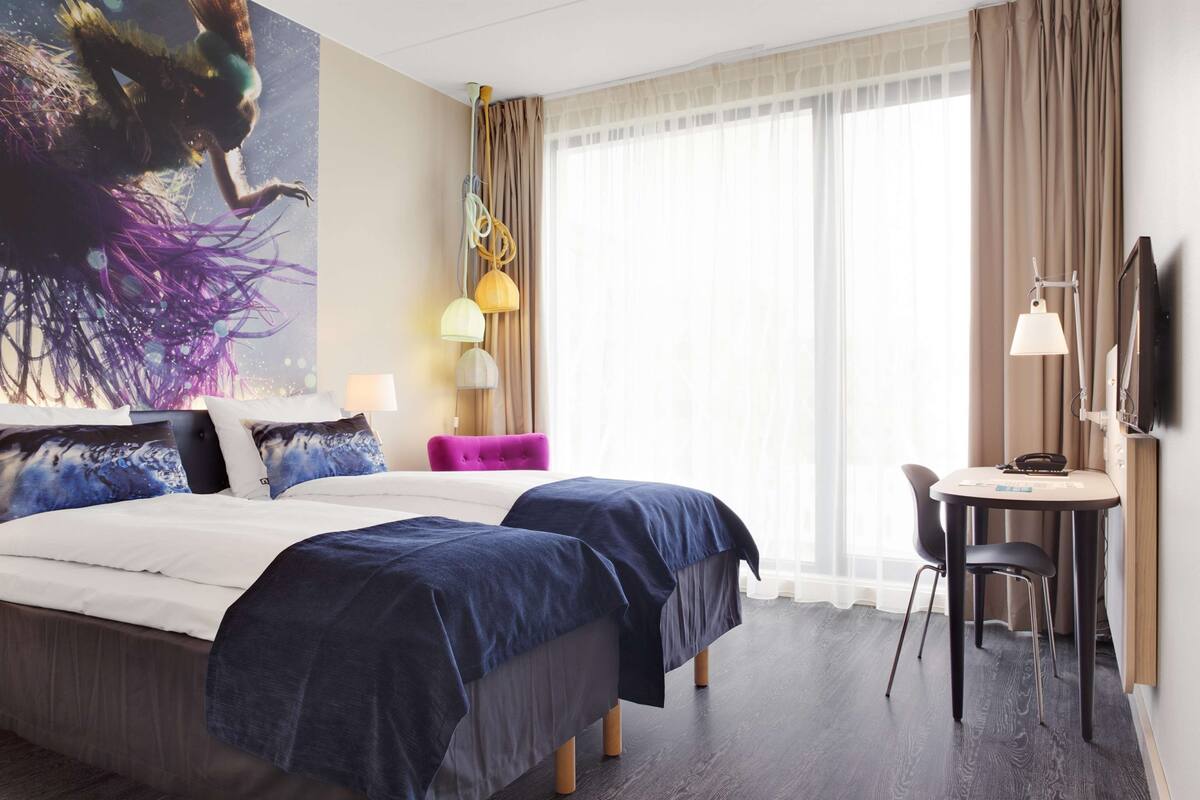 Habitación de hotel con cama, dos mesitas de noche, un escritorio con silla, una ventana grande con cortinas y un televisor montado en la pared.