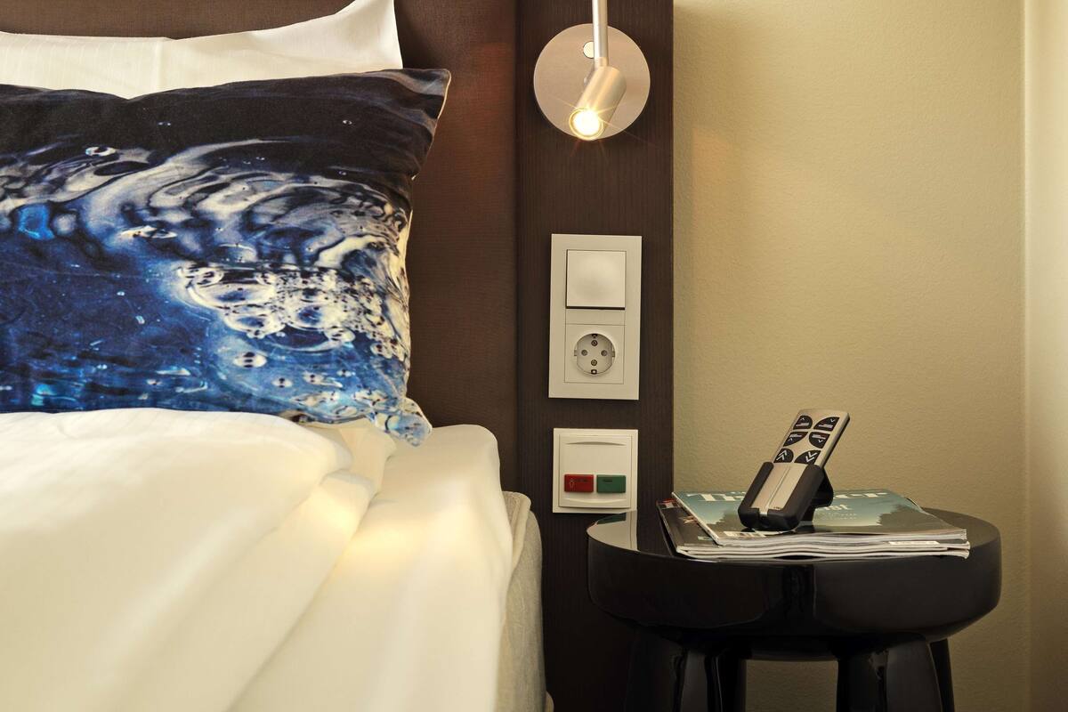 Una habitación de hotel con una cama, una mesita de noche con un control remoto y una lámpara fijada a la pared.
