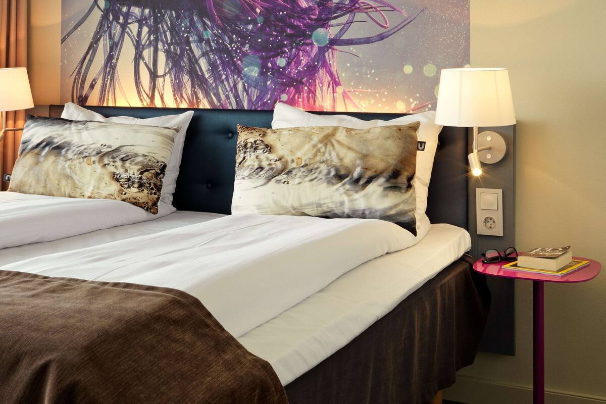 Habitación de hotel con cama, dos almohadas, mesita de noche con lámpara y una obra de arte en la pared.