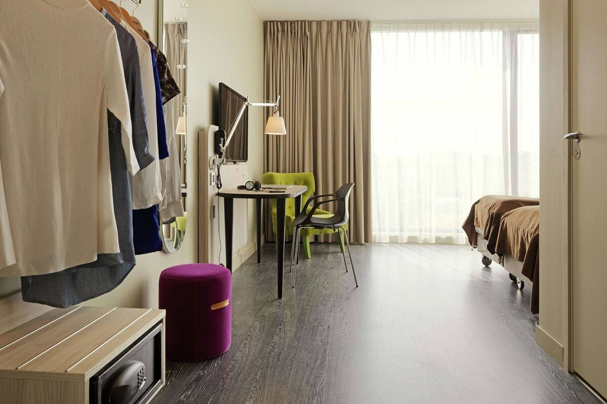 Habitación de hotel con cama, escritorio con lámpara, silla, mesita, armario y ventana con cortinas.