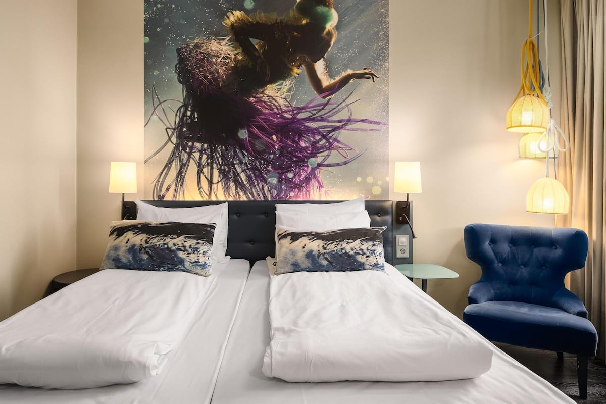 Habitación de hotel con una cama grande, dos almohadas, un sillón azul, una mesita y una obra de arte en la pared.