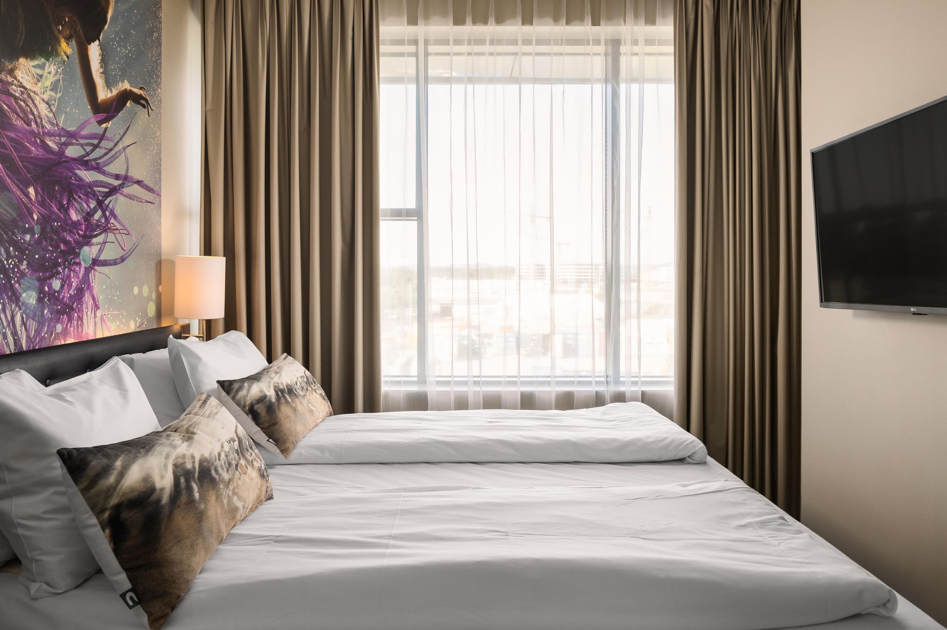 A hotel room with a large bed, a flat-screen TV, a window with curtains, and a painting on the wall.