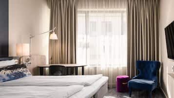 A hotel room with a bed, a desk, a chair, a lamp, and a window with curtains.
