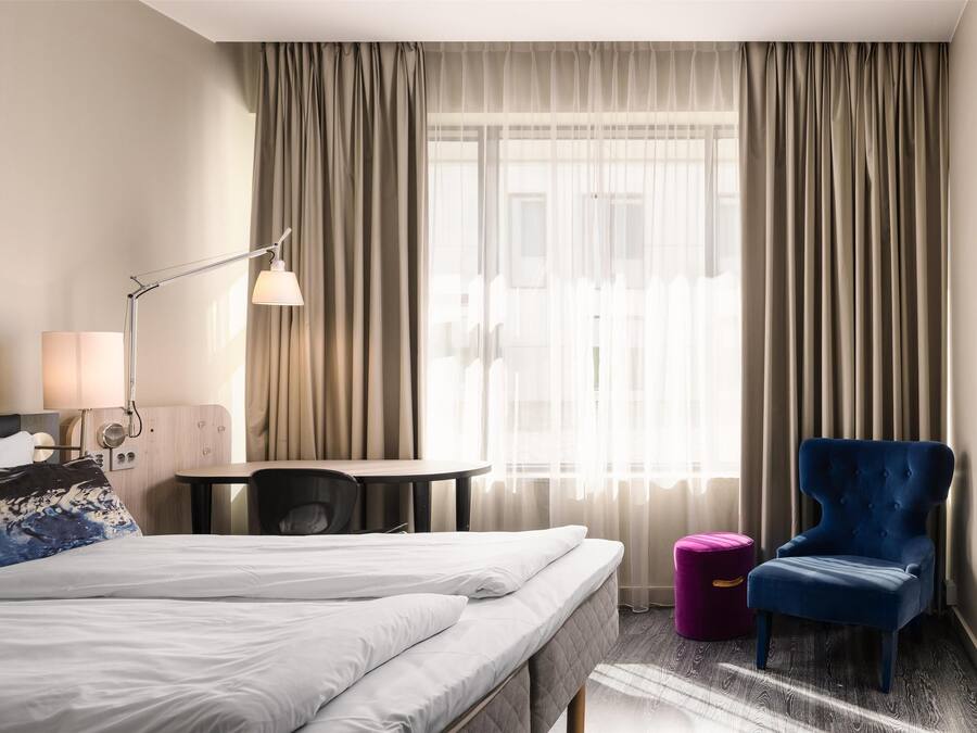 A hotel room with a bed, a desk, a chair, a lamp, and a window with curtains.
