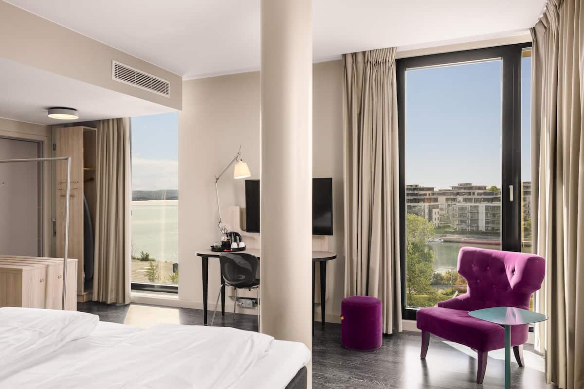 Habitación de hotel con cama, escritorio, silla, televisor y ventana grande con vista a la ciudad.