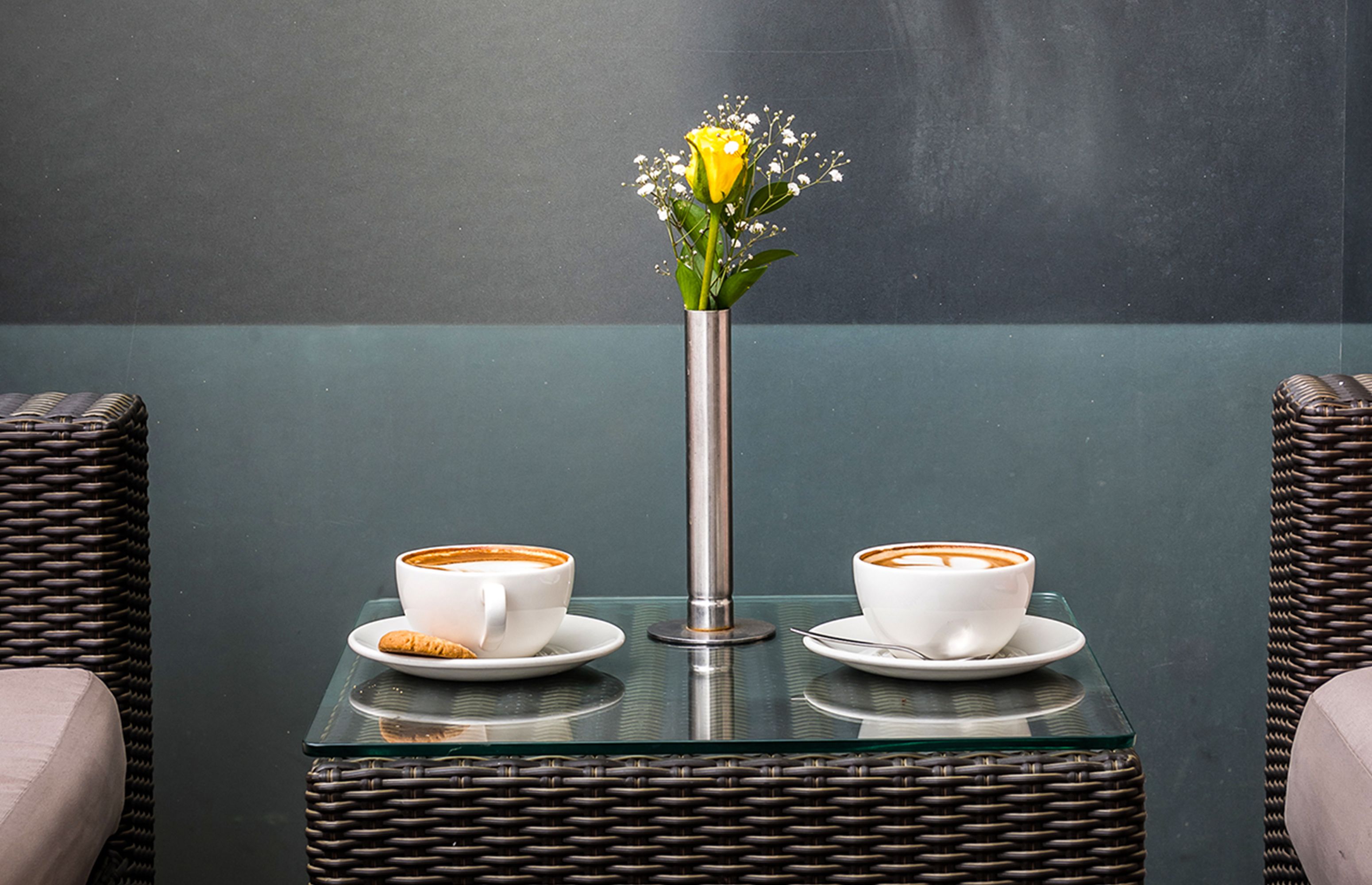 Una mesa con dos tazas blancas de café y un florero con una flor amarilla.