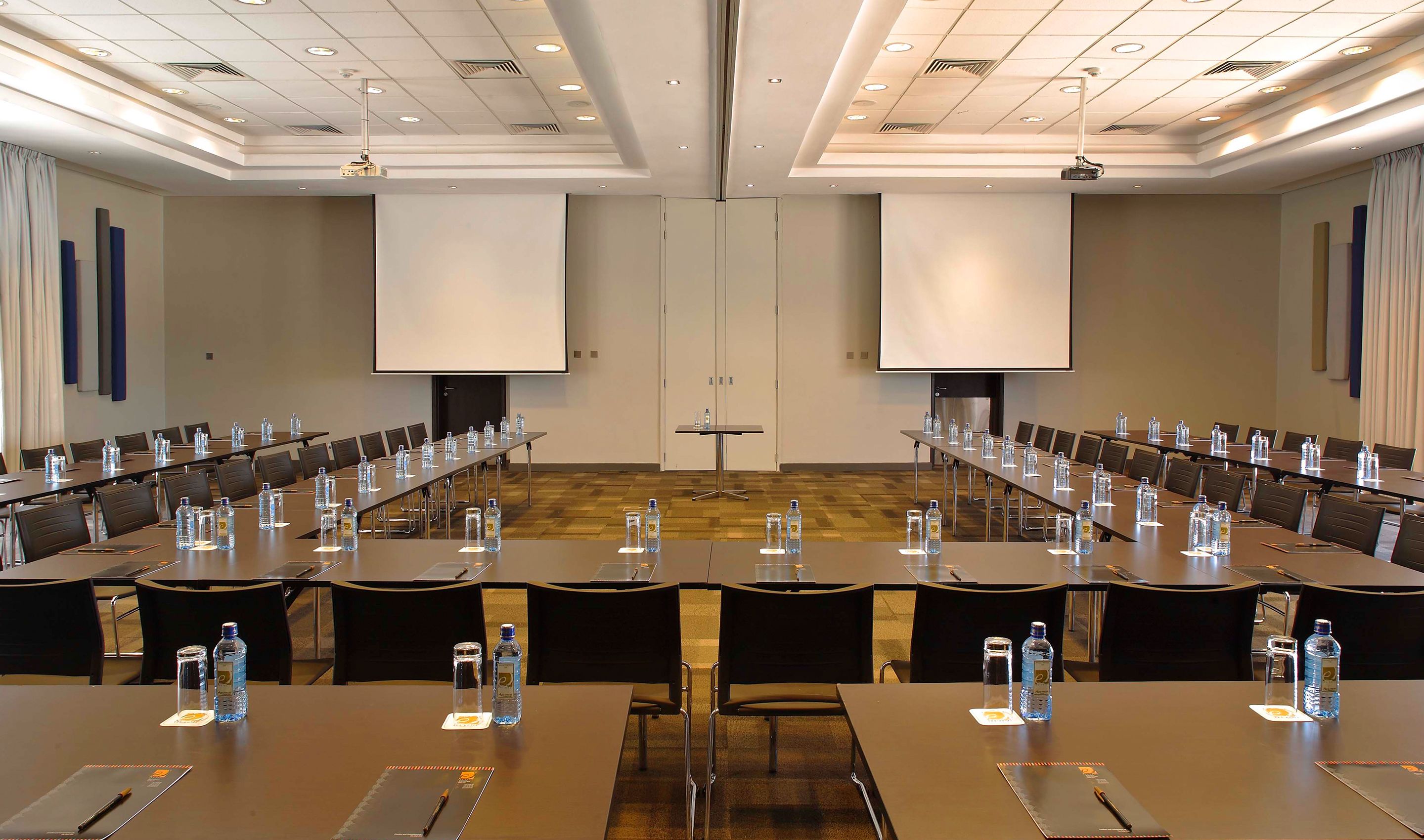 Une salle de conférence équipée de longues tables, de chaises, de bouteilles d’eau et d’un écran de projection.