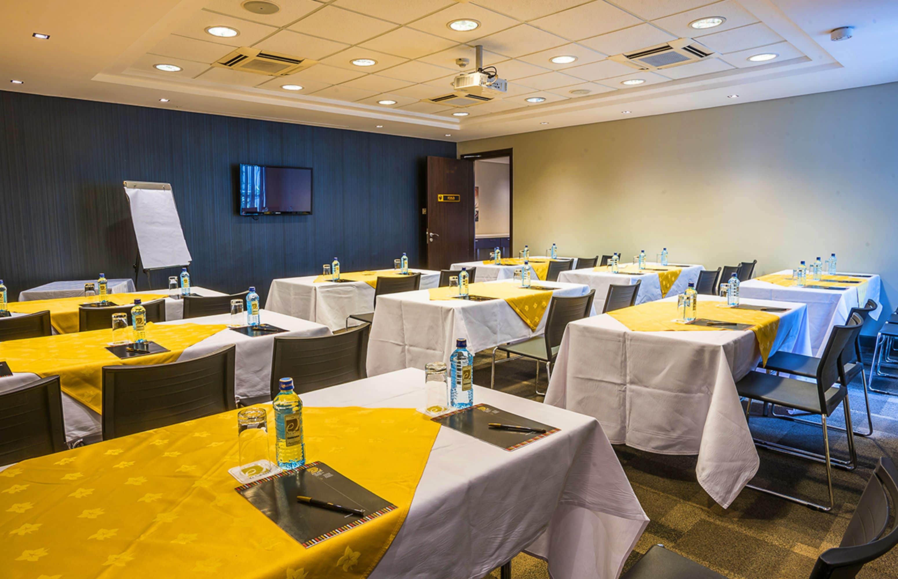 Ein Konferenzraum mit mehreren Tischen, jeder mit einer gelben und weißen Tischdecke bedeckt, ausgestattet mit Wasserflaschen, Gläsern und Notizblöcken.