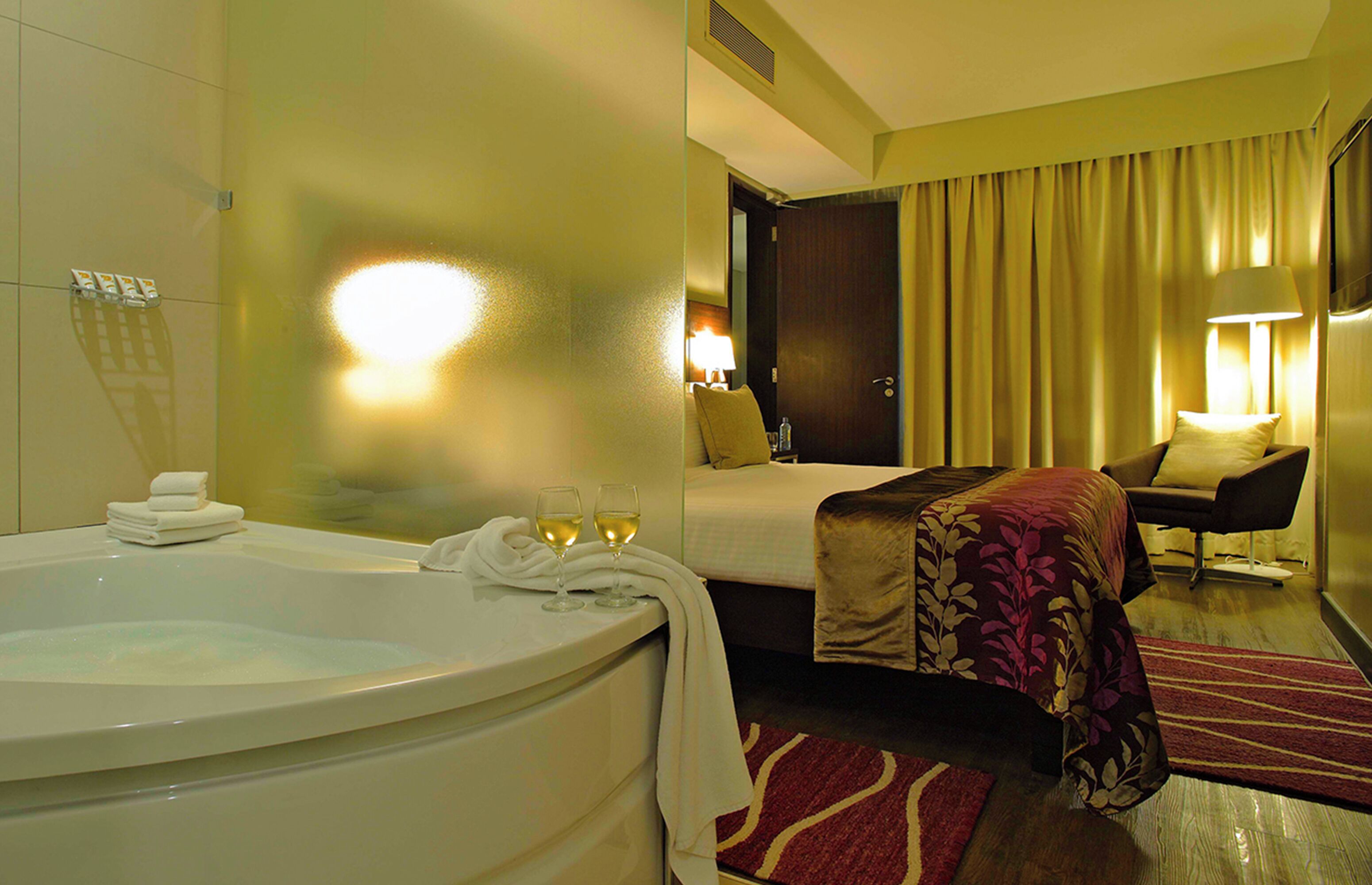Habitación de hotel con una bañera grande, una cama con cubrecama rojo y negro, una zona de estar con una silla y una mesa, y una puerta que da a otra habitación.