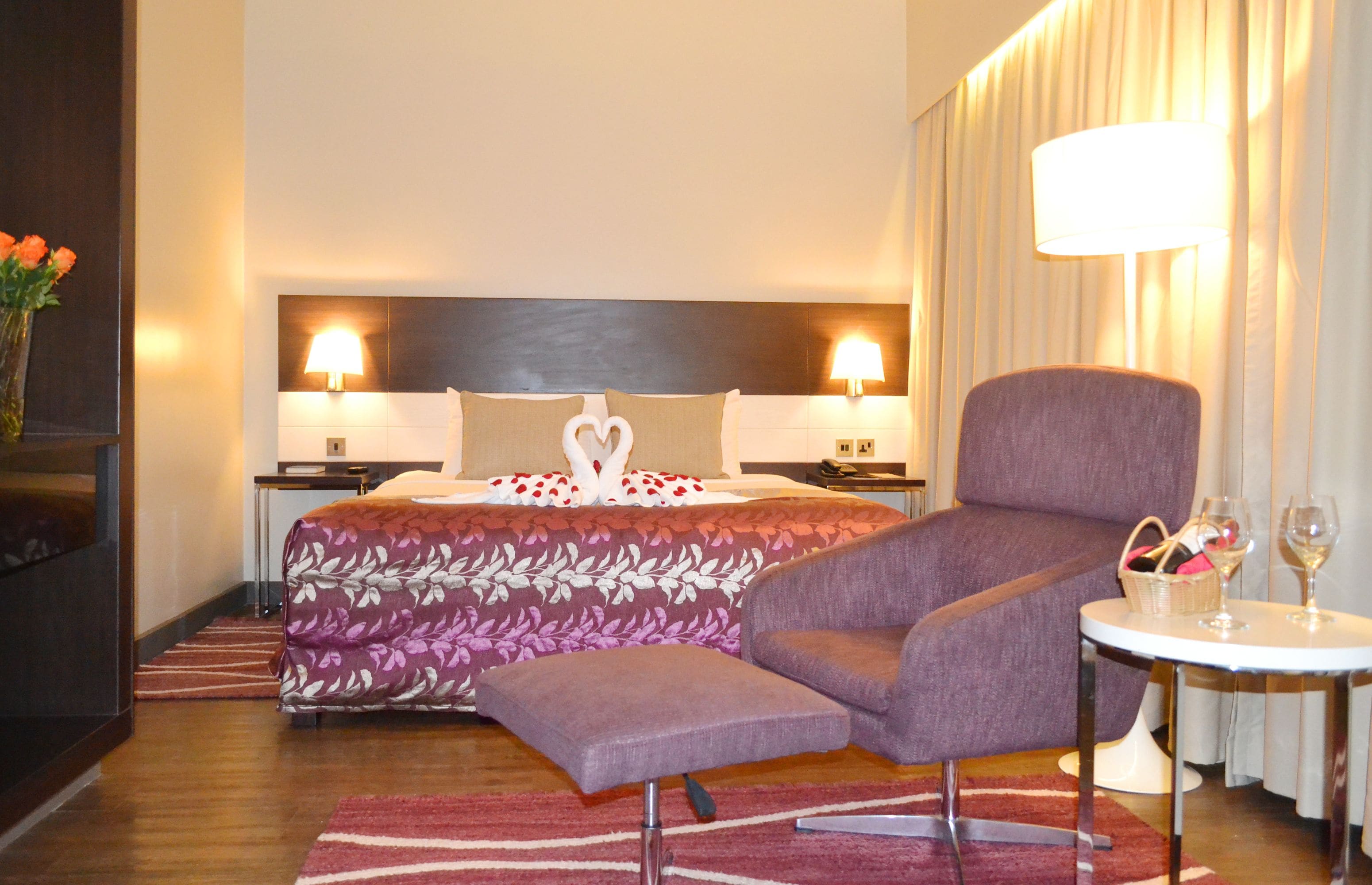 Habitación de hotel con cama, dos lámparas de noche, un sillón púrpura, una mesita con una copa de vino y un jarrón con flores.