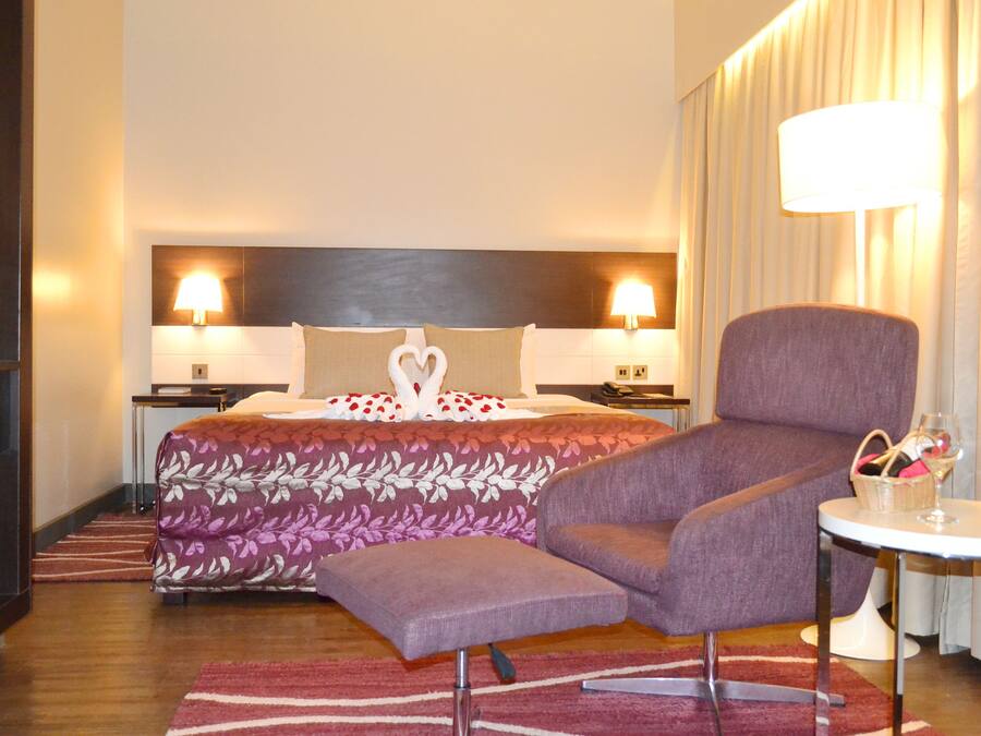 Habitación de hotel con cama, dos lámparas de noche, un sillón púrpura, una mesita con una copa de vino y un jarrón con flores.