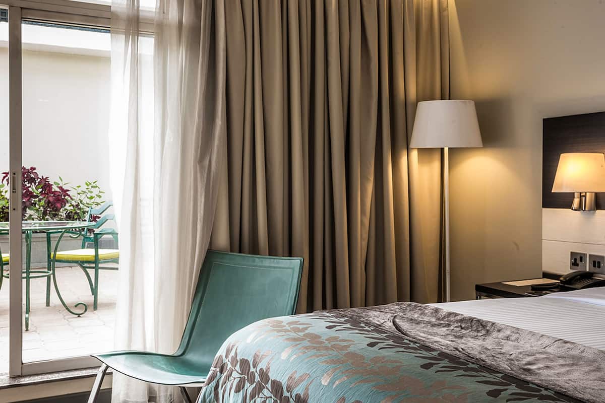 A hotel room with a bed, a chair, a table with plants, a lamp, and a view of the outdoors.