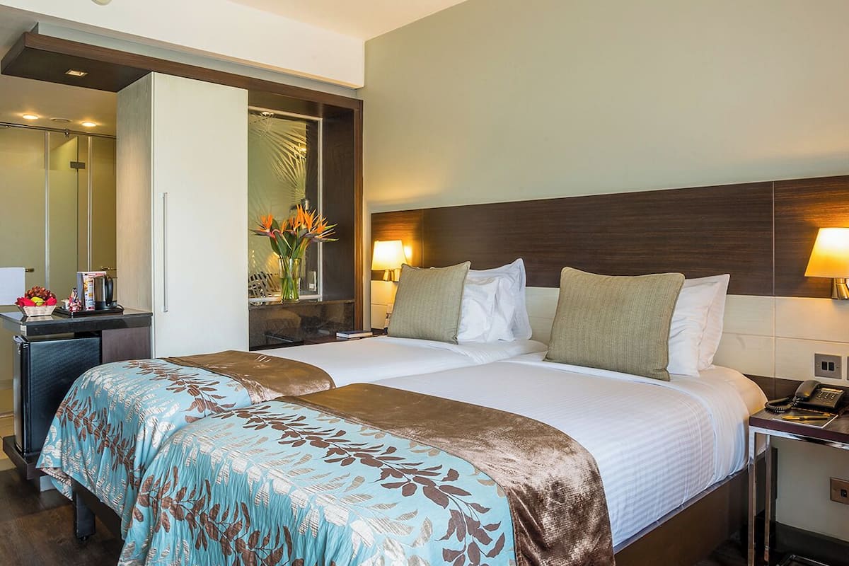 A hotel room with two beds, a desk, a mirror, and a nightstand with a lamp.