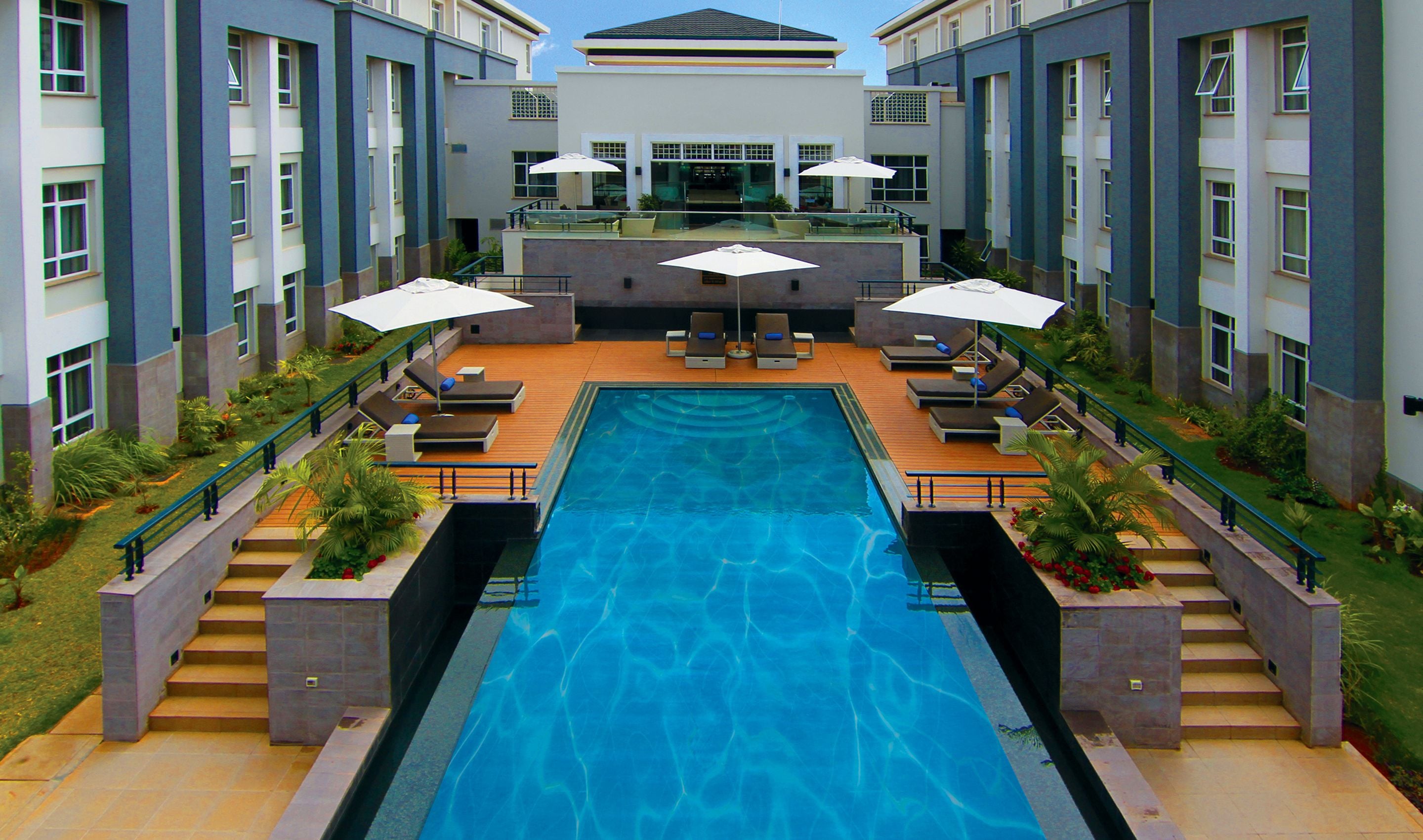 A swimming pool with lounge chairs and umbrellas, surrounded by a building with multiple floors and balconies.