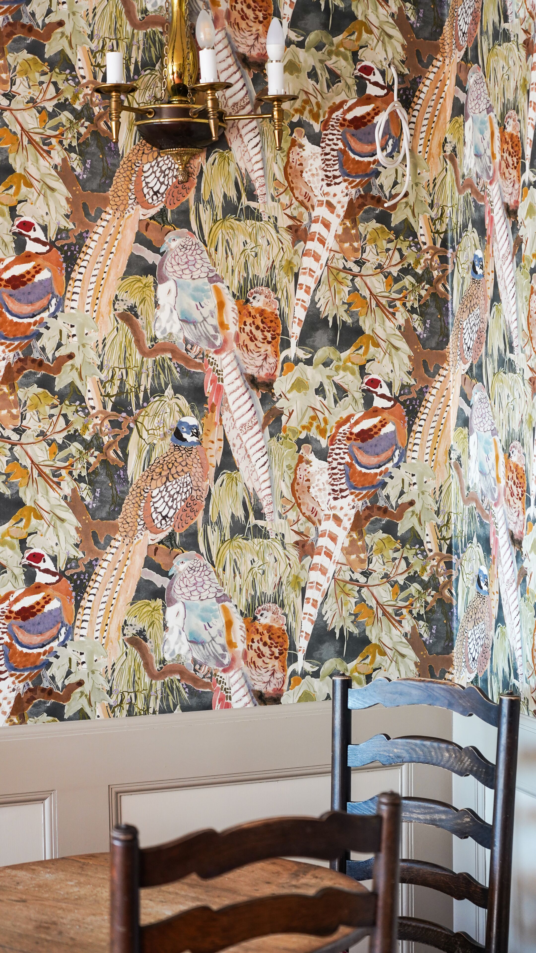 A dining area with a table and chairs, a wall with a detailed mural of birds and foliage, and a chandelier with multiple light fixtures.