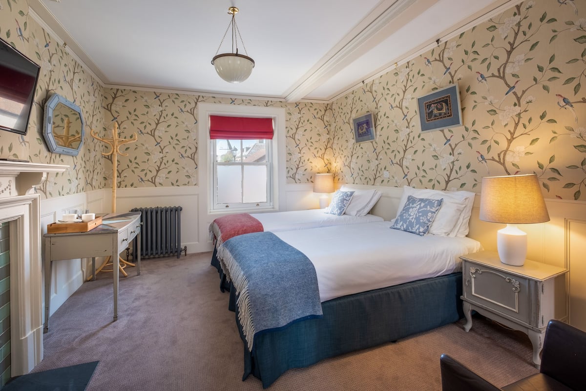 A bedroom with a double bed, bedside lamps, a dresser, a mirror, a radiator, and a window with a red valance.