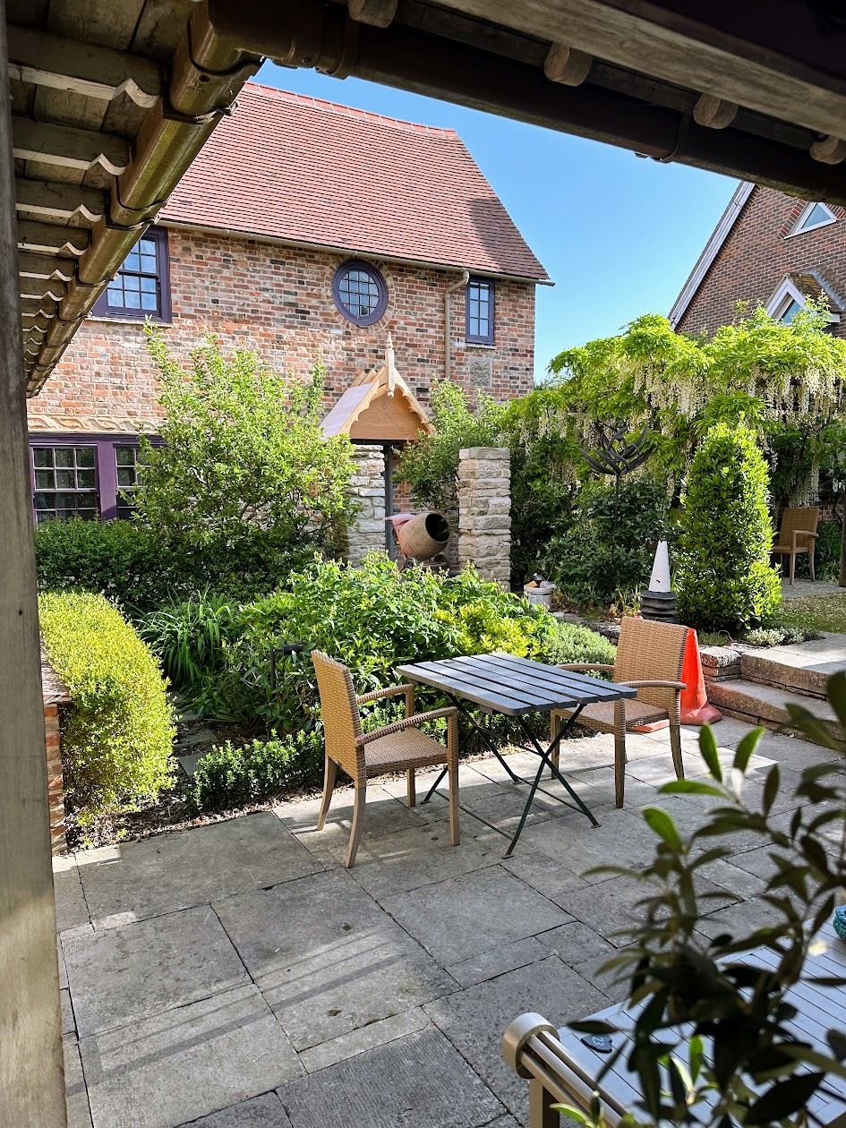 A patio area with a table and chairs, surrounded by greenery and a brick building in the background.